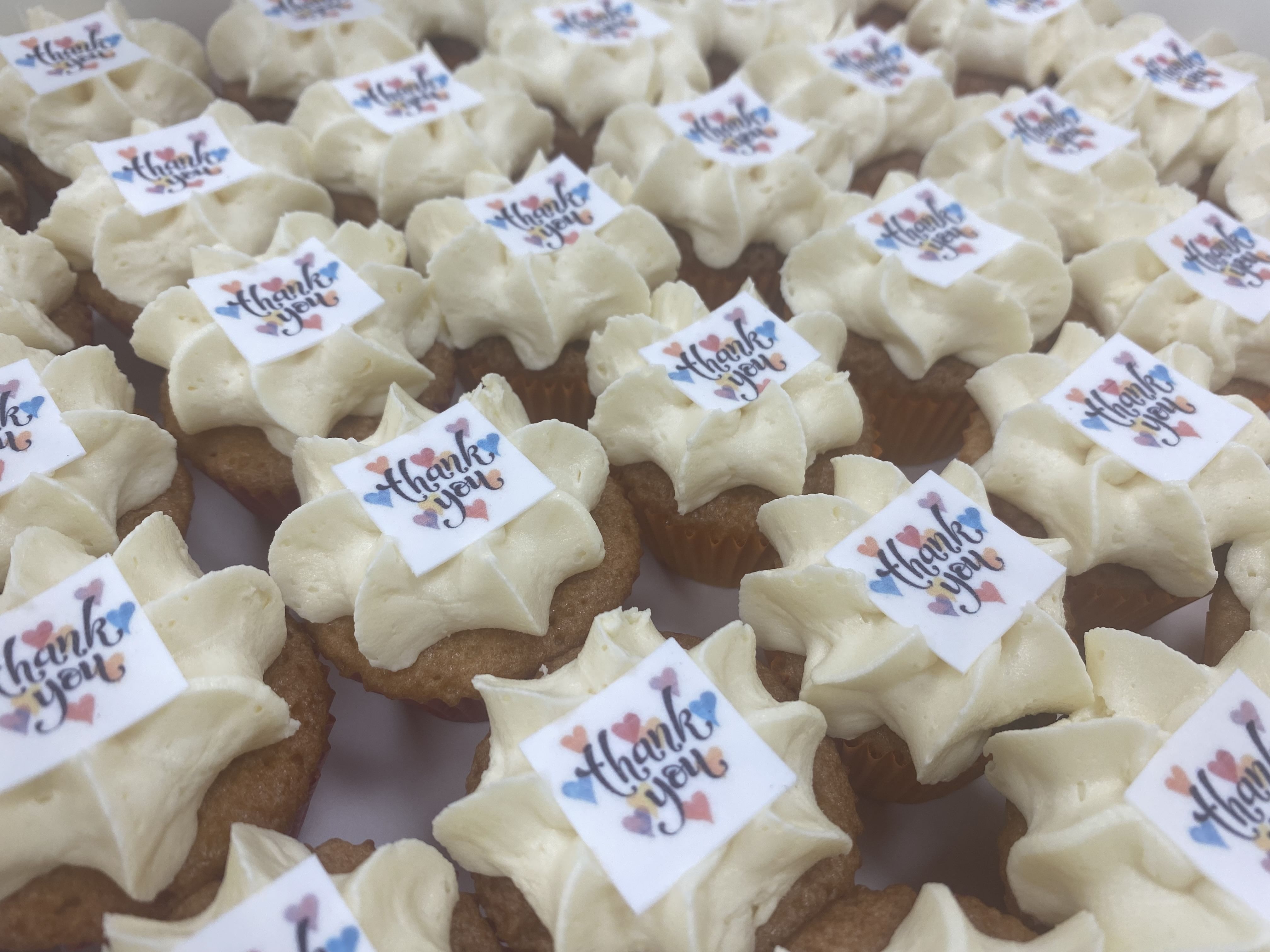 A tray of mini vanilla cupcakes topped with white frosting and edible decorations that read 'Thank you' in colorful script. Perfect for corporate events, the cupcakes are arranged in neat rows, each featuring a small rectangular label on top.